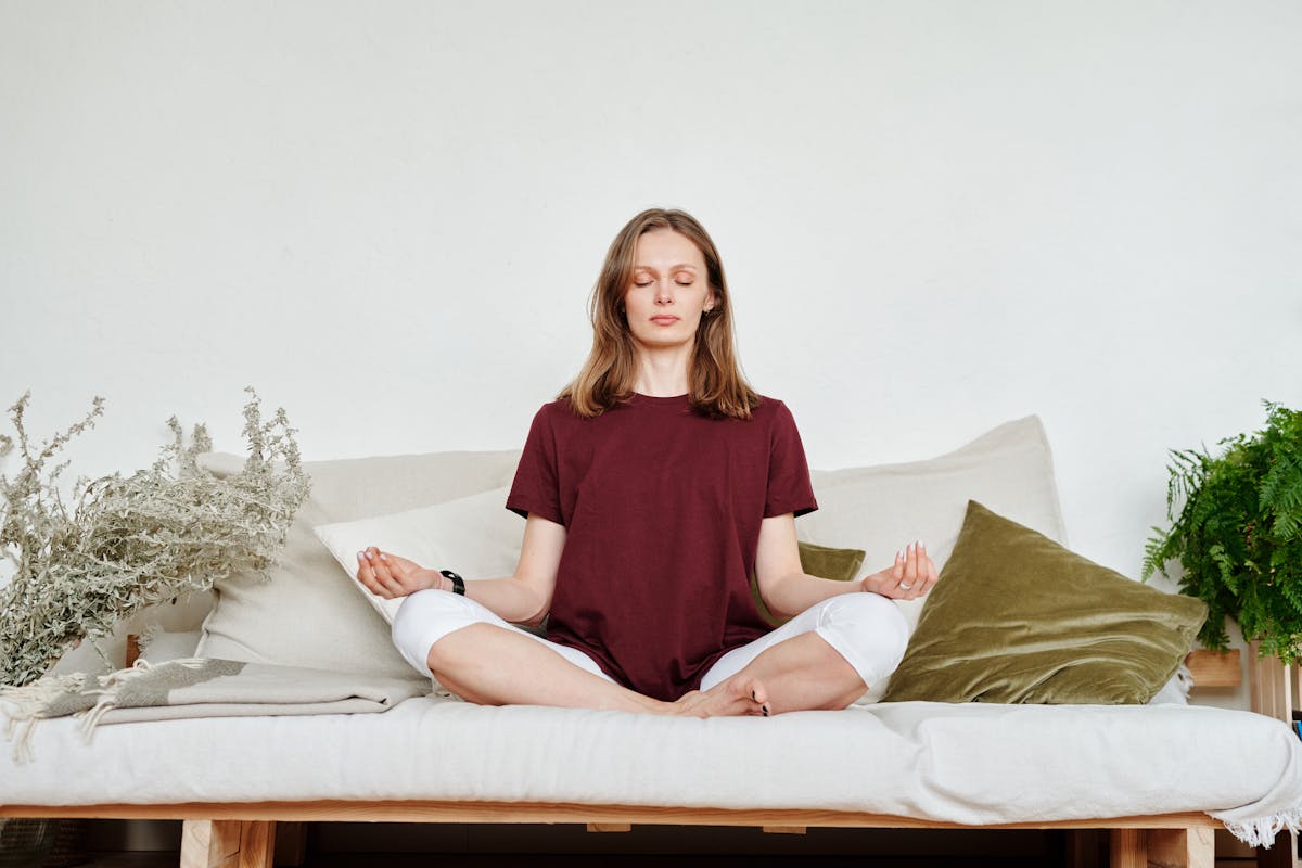 A woman sitting in meditation on a sofa surrounded by indoor plants, eyes closed in calm focus