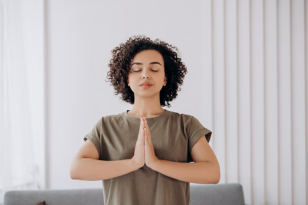 A woman with curly hair sitting cross-legged in meditation, eyes closed, in a calm indoor space